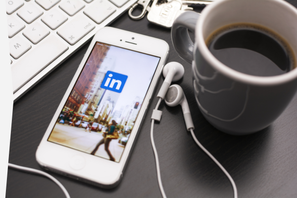 A smartphone displaying the LinkedIn app sits on a desk beside earphones, a keyboard, keys, and a cup of coffee.