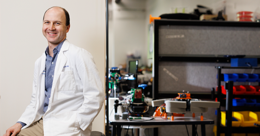 A physician in a white coat sits smiling beside a lab workspace filled with robotics and engineering equipment.