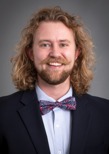 Mark Schmidt smiling a dark suit jacket and patterned bow tie against a gray background.