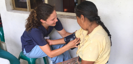 Healthcare worker in scrubs taking blood pressure of a seated person in a yellow shirt and patterned skirt outdoors near a barred window.