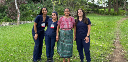 Four people stand outdoors, three in blue medical scrubs and one in traditional clothing, smiling together.