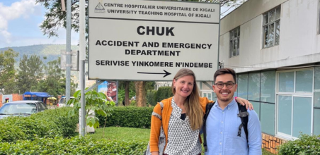 Two people standing in front of the Accident and Emergency Department sign at Kigali University Teaching Hospital, with hospital building and hills in the background.