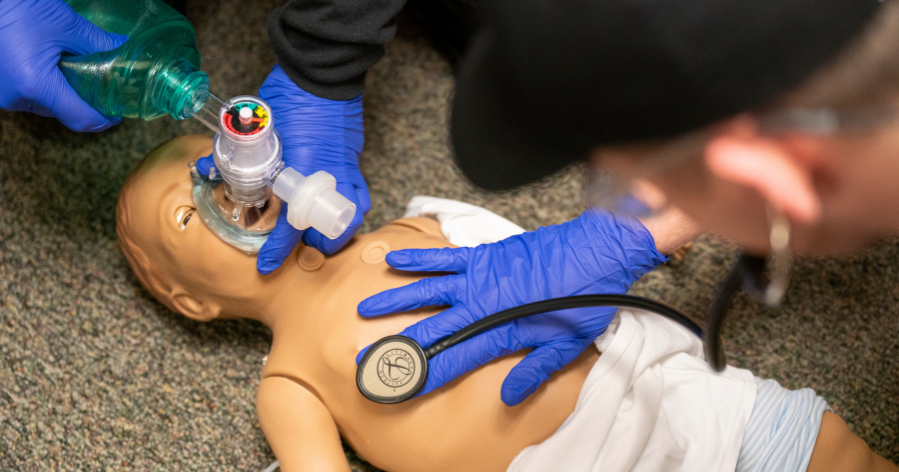 Gloved hands provide CPR to an infant manikin using a bag-valve mask while listening with a stethoscope.