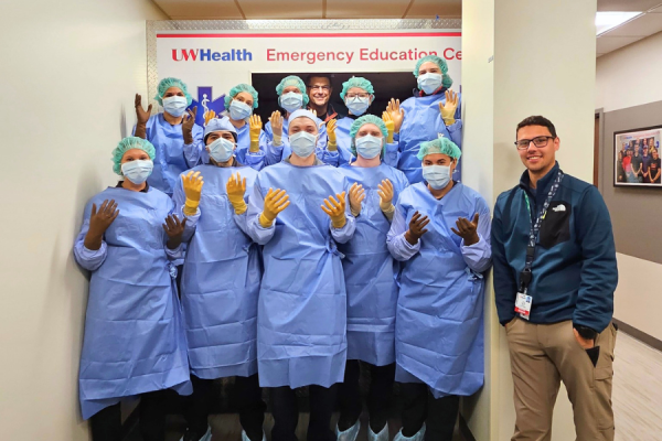 Group of healthcare trainees in surgical gowns, masks, and gloves pose in the UW Health Emergency Education Center with an instructor nearby.