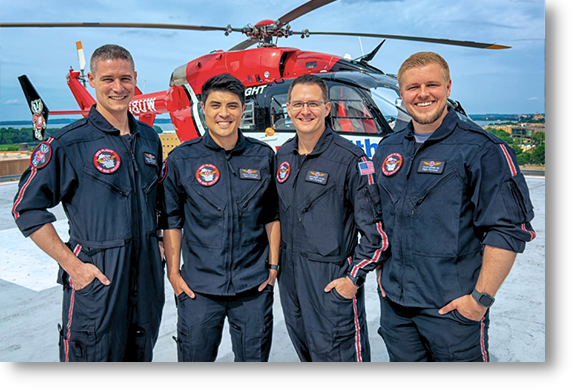 Four Med Flight crew members in black flight suits stand together smiling on a rooftop helipad overlooking the city.