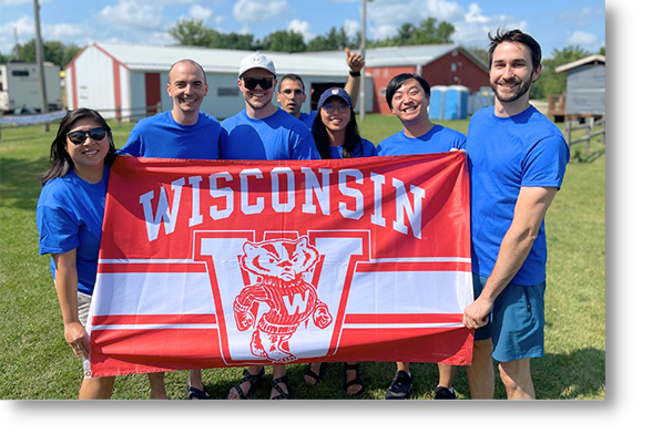 Seven people hold a University of Wisconsin flag outdoors in a grassy field.