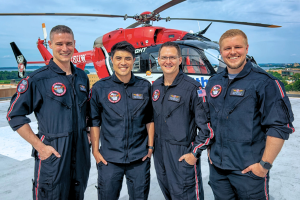 Four Med Flight crew members in black flight suits stand together smiling on a rooftop helipad overlooking the city.